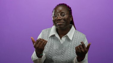 Gözlüklü ve gri süveterli, mor arka planda elleri havada gülümseyen Afrikalı Amerikalı bir kadın..