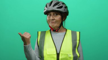 Yeşil arka plana karşı miğfer ve güvenlik yeleği giyerken başparmağıyla işaret eden kadın, güvenli açık hava faaliyetlerine güven ve güvenin bir göstergesi..