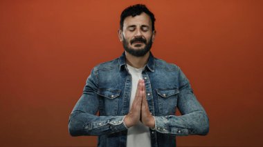 İspanyol bir adam turuncu bir arka plan karşısında sakal ve kot ceketle meditasyon yapıyor. Dua eden eller, gözleri kapalı, sükunet ve farkındalık sergiliyor.