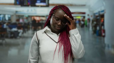 Saçları örgülü bir kadın havaalanı terminalinde gözlük takıyor. Gezginlerle dolu bulanık bir arka plana karşı düşünceli görünüyor..