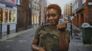 Kamuflaj üniforması giyen genç Afrikalı Amerikalı kadın asker yağmurlu bir şehir caddesinde zafer işareti yapıyor:.