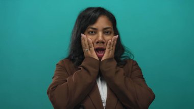 Kahverengi ceketli kadın, elleriyle yanaklarını tutuyor ve turkuaz stüdyo duvarına karşı ağzını açıyor. Sürpriz şok..