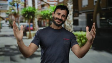 Sakallı İspanyol bir adam şehir caddesinde ağaçlar ve binalar arkasında mutluluk ve kendine güveni ifade ederek neşeli bir şekilde dolaşır..