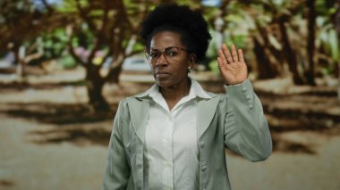 Parkta elini kaldıran gözlüklü ve ceketli bir kadın güneşli bir günde ağaçlarla çevrili doğal bir açık hava ortamında güç ve güveni simgeler..