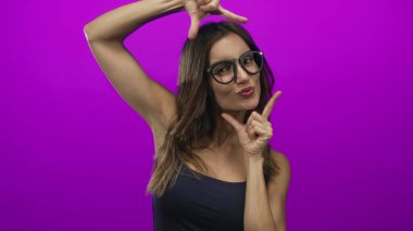 Gözlüklü bir kadın sıcak ışık altında gülümser ve yüzünü mor bir stüdyoda çerçevelemek için çıplak ellerini kullanır..