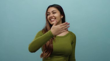 Mavi arka planda yeşil bluzlu gülümseyen kadın izole bir ortamda mutluluk işareti yapıyor neşeli İspanyol ifadesi ve pozitif titreşimler sergiliyor.