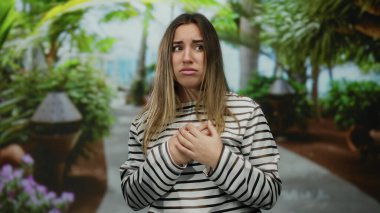 Çizgili tişörtlü genç bir kadın açık havada, yemyeşil bir parkta yüzünü örterek doğanın ortasında huzursuzluk ve endişe dolu duygular taşıyor.