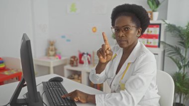 Beyaz önlüklü kadın doktor pediatri kliniğinde klavyeye yazarken kameraya işaret parmağıyla işaret ediyor; otorite bakımı.