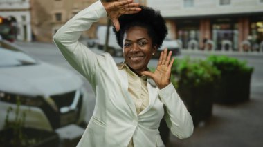 Beyaz ceketle gülümseyen bir kadın şehrin arka planında jestler yaparak öğleden sonraki şehir sahnesinde neşe ve kendine güveni somutlaştırıyor..