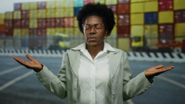 Kargo konteynırlarının önünde açık havada duran, kafası karışmış bir ifade ve kolları açık bir kadın, hareketli sahil ortamında belirsiz bir an sergiliyor..