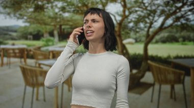 Kadın gergin el hareketleriyle akıllı telefonu kulağına tutuyor. Görünür bir buruşukluk ve çatık kaşlar binanın içinde. Hayal kırıklığı..