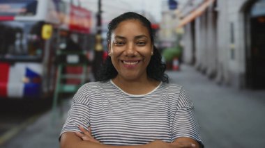 Beyaz çizgili tişörtlü kadın, bulanık binalarla dolu hareketli bir caddede kolları çapraz duran kırmızı bir otobüste duruyor..