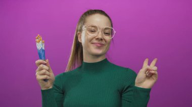 Kadın stüdyo ortamında birkaç boya fırçası tutarken, gözlük ve yeşil balıkçı yaka ile gülümsüyor; yaratıcı neşe.