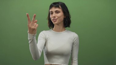Kısa siyah saçlı, beyaz bir bluz giyen ve stüdyoda parmaklarını gösteren Latin kökenli genç bir kadın..