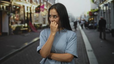 Bir şehir caddesinde tırnaklarını yiyen genç İspanyol kadın, el ele ve kollar çapraz; endişe belirsizliği.