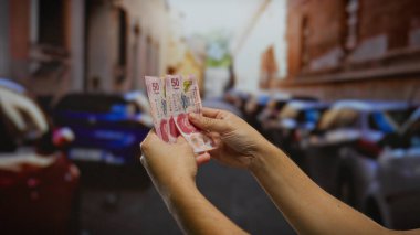 Meksika pesolarını işlek bir şehir caddesinde park edilmiş arabalarla tutan adam, finans, bankacılık ve şehir hayatı temaları, iş ve yatırım görselleri için ideal.