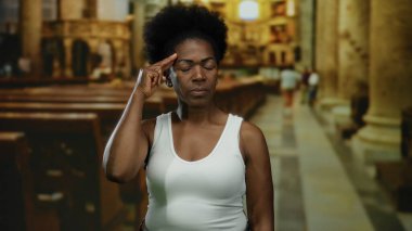 Kilisede duran bir kadın, arka planda bulanıklığa karşı düşünceli bir ifadeyle kafasına dokunuyor, beyaz atlet giyip iç mekan ayarları yapıyor..