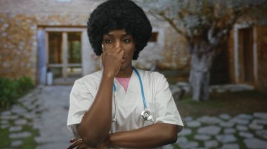 Steteskoplu genç Afrikalı Amerikalı kadın doktor bina girişinde burnunu çimdikliyor..