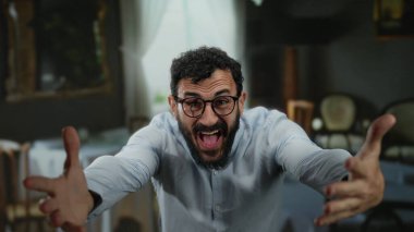 Sakallı ve gözlüklü İspanyol bir adam boş masa ve sandalyelerle çevrili rahat bir restoranda neşeli hareketler yaparak davetkar bir atmosfer yaratıyor..