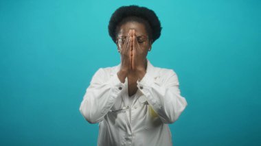 Beyaz önlüklü ve steteskoplu kadın doktor, gözlük takıyor, ellerini yüzüne koyuyor sonra da avuçlarını turkuvaz stüdyosuna karşı açıyor..