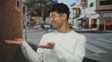 Genç İspanyol gülümseyen bir adam, kalabalık bir şehir caddesinde, sıcak bir davet sunum jesti avuç içlerini uzatır.