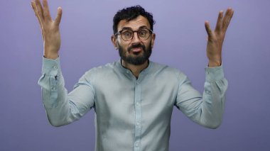 Mor bir arka plana karşı çılgın ve karışık bir ifade sergileyen sakallı İspanyol bir adam, gözlük takıyor ve açık mavi gömlek giyiyor..