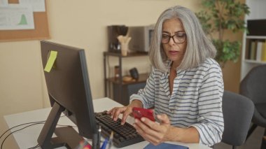 Bir ofis binasında kırmızı bir akıllı telefonu elinde tutarken elleri klavyede yazan bir kadın; konsantrasyon verimliliği.