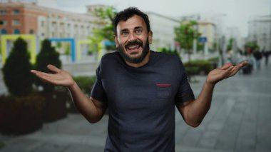 Arka planda bulanık binalar olan bir sokakta belirsizlik gösteren sakallı İspanyol bir adam..