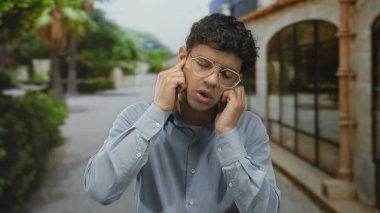 İnsanlar, ağaçlarla ve yeşilliklerle dolu güneşli bir sokakta dikilirken kulaklarını parmaklarıyla kapatırlar..