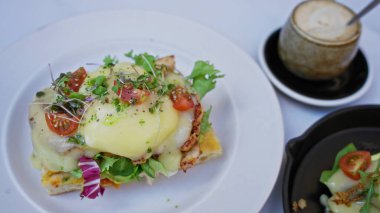 Taze yeşillik ve domatesli yumurtalar kapuçinonun yanında beyaz bir tabakta duruyor. Kafeterya masasında yakalanmış, canlı kahvaltı lezzetlerini vurguluyor..