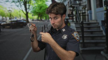 Polis üniformalı bir adam, elinde kelepçeler ve bir şehir caddesinde park etmiş arabaların yanında kamerayı işaret ediyor..