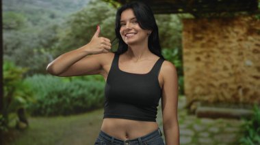 Siyah ekin bluzlu ve kot pantolonlu kadın yeşil ormanda taş bina yolunun açıklığında baş parmak hareketi yapıyor; iyimserlik.