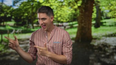 Yeşil ağaçların arasında parktaki iki parmağı gösteren adam, gülümseyen ve öne doğru eğilen; oyuncu özgüven.