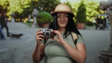 Sunny Park 'ta elinde kamera olan bir kadın. Şapka takıyor ve ağaçlarla gülümsüyor.