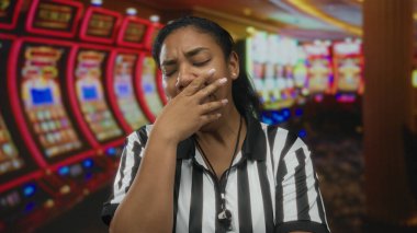 Kumarhane binasında çizgili gömlek giyen Afrikalı Amerikalı kadın hakem ağzını slot makinesiyle kapatıyor..