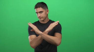 Genç İspanyol asıllı bir adam, yeşil bir stüdyo arka planında x jesti yaparak kollarını kavuşturuyor, kameraya doğru sert bir ifadeyle bakıyor;.