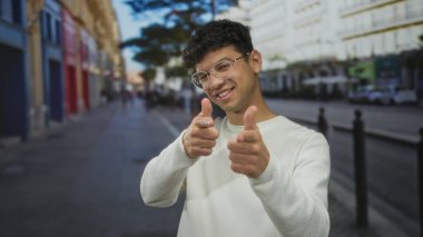 Tel çerçeveli gözlük takan bir adam güneş ışığıyla aydınlatılmış şehir caddesinde silah hareketi yapıyor. Binalarla dolu..