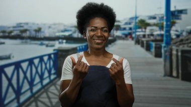 Önlüklü Afrikalı Amerikalı bir kadın davetkar bir şekilde deniz kenarında bir gezinti yapar, arka planda deniz ve plaj vardır..