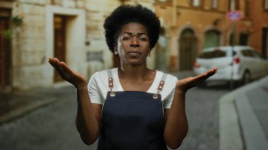 Önlüklü kadın, umut dolu bir jestle şehir caddesinde duruyor. Yumuşak ışık altında sakin bir ortam yaratıyor. Derin bir düşünce ve samimiyet..