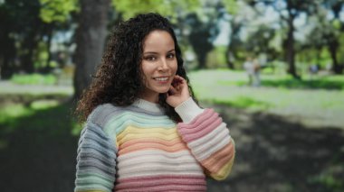 Açık havada güneşli bir günde arka planda ağaçlarla parkın tadını çıkaran renkli kazaklı bir kadın..