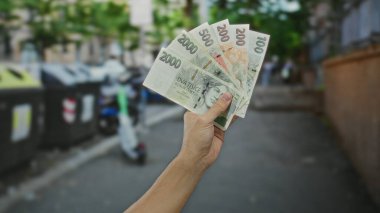 Geri dönüşüm kutularının yakınındaki bir sokakta, para ve çevre odaklı şehir merkezinde beyaz bir erkek resmedilmiş, el ele tutuşan koruna banknotları..