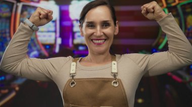 Kumarhanede kaslarını esneten genç bir kadın kahverengi önlük giyiyor ve arka planda kumar makineleri var..