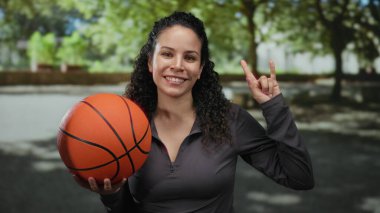 Genç İspanyol kadın elinde basketbol topuyla açık hava parkında jest yapıyor. Ağaçlarla çevrili güneşli gökyüzünün altında enerji ve coşku gösterisi yapıyor..