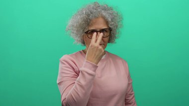 Yeşil arka plana karşı ihtiyatlı davranan, gözlük takan ve pembe kazak giyen gri saçlı yaşlı kadın, sosyal farkındalığa ilgi çekici bir odak katıyor..