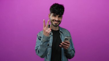 Akıllı telefonuyla gülümseyen genç adam izole edilmiş pembe arka planda kot ceket giyiyor.