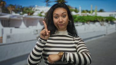 Kadın parmağı beyaz bina ve palmiye ağaçları ile sokakta bileğindeki saati işaret ediyor, çizgili üst görünürde; sabırsızlık dakikliği.