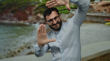 Sahilde sakallı ve gözlüklü bir adam ellerini deniz manzarasını çerçeveletip doğada yaratıcılık ve neşe ifade ediyor..
