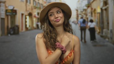 Turuncu bikinili ve geniş hasır şapkalı kadın, güneş ışığı altındaki bir kaldırım taşı caddesinde sessizlik için parmaklarını dudaklarına koyar..