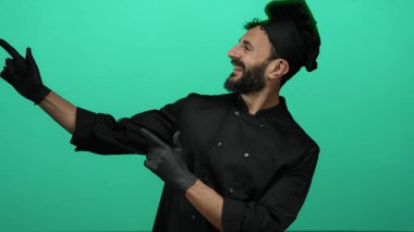 Sakallı İspanyol erkek aşçı canlı yeşil bir fon karşısında şakacı bir şekilde yan yatmış, siyah şef kıyafeti ve uyumlu bir şapka giymiş, aşçılık hevesini somutlaştırıyor..