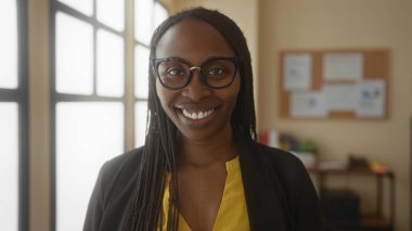 Ofis ortamında gözlüklü gülümseyen, parlak camlı, sarı gömlekli, kıvırcık saçlı, kendine güveni ve profesyonelliği olan bir kadın..